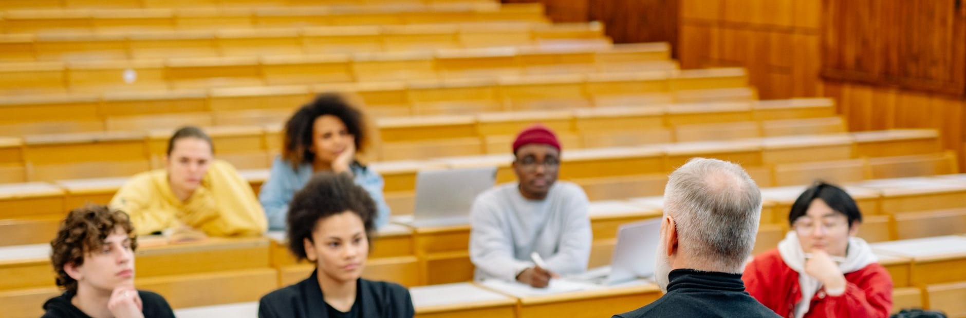 Six students spread thinly across the tiered wooden benches of a lecture theatre, most of the rows behind them empty, facing a grey-haired instructor seen from behind