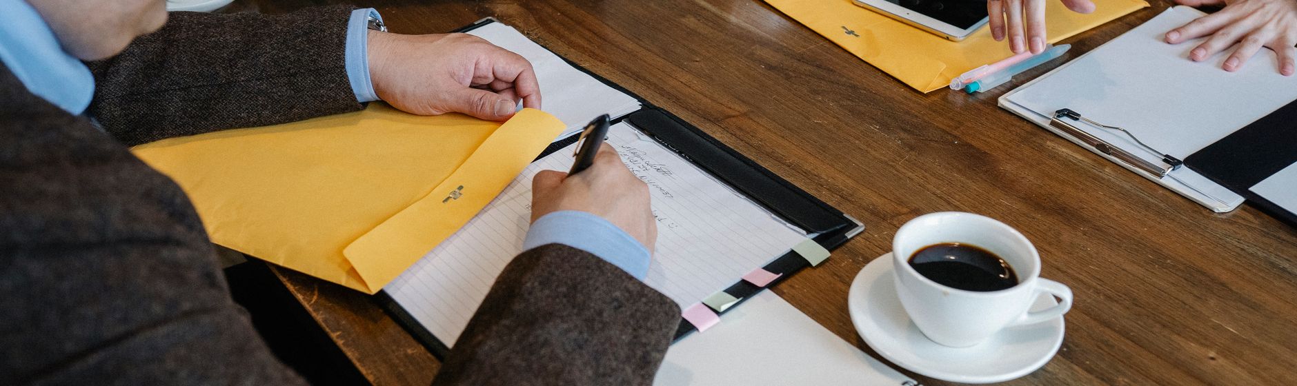 Two people work across a wooden table, one writing on a tabbed legal pad next to a pair of yellow envelopes and the other resting a hand on a clipboard, with a white cup of black coffee at the edge of the frame