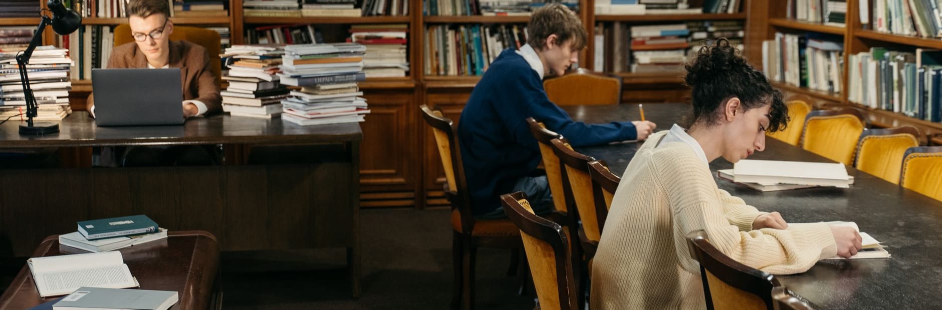 Three people work at long dark wooden tables in a library reading room, one behind a laptop and a stack of books, the others bent over open volumes, with filled shelves running along the wall behind them