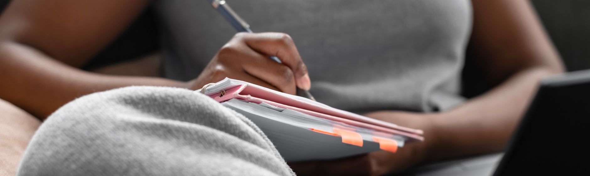 Close-up of a person sitting with a spiral notebook and tabbed papers resting on their lap, pen in hand, a dark tablet propped beside them