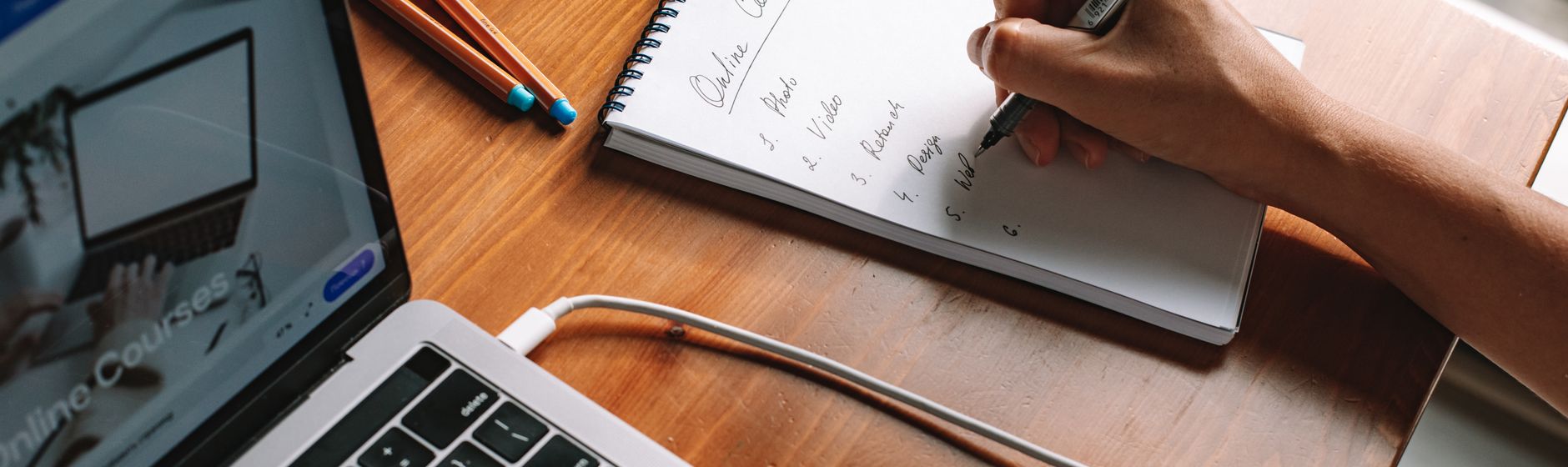 A hand writes a numbered list in a spiral notepad on a wooden desk beside an open laptop showing an online courses page, with two marker pens and a charging cable alongside