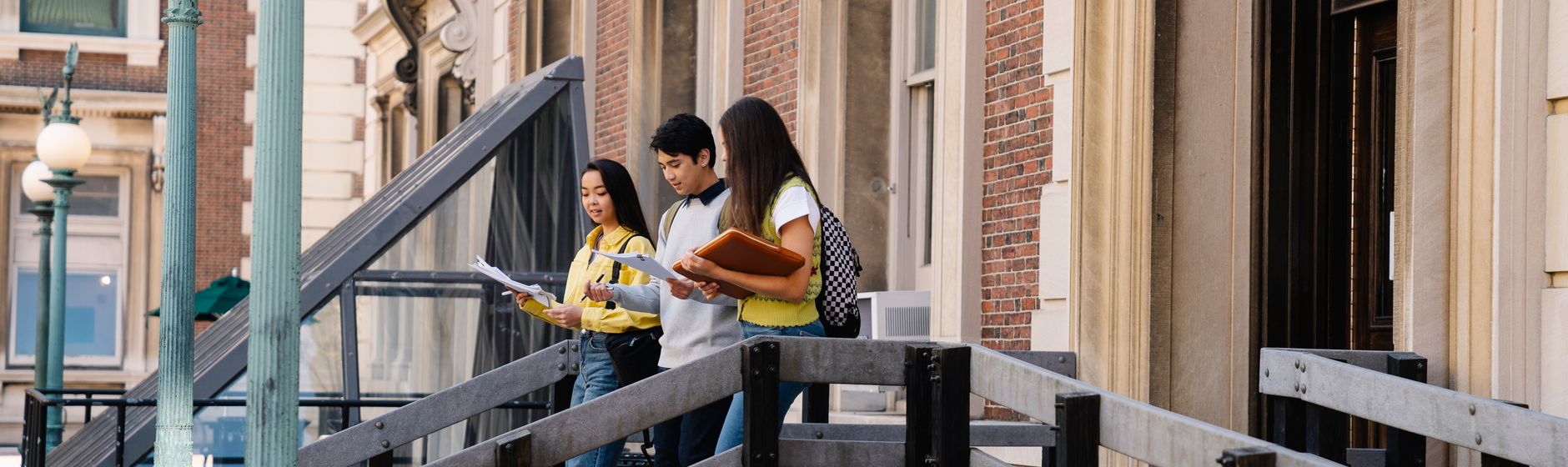 Three students review papers together on the steps of a brick university building